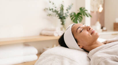 Person relaxing in a spa, eyes closed, with a white headband on and a towel-covered table.