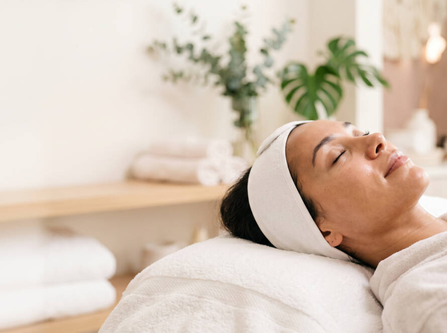 Person relaxing in a spa, eyes closed, with a white headband on and a towel-covered table.